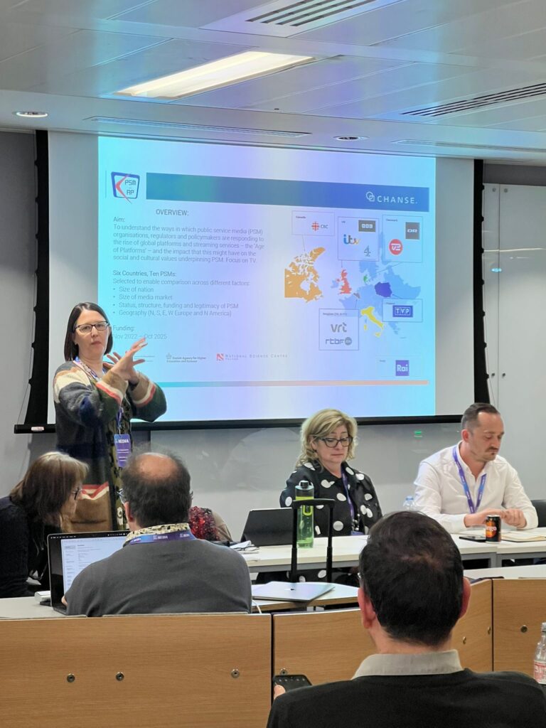 Catherine Johnson is standing infront of a projection presenting on the PSM-AP project. The screen displays a slide with an overview of the project. There are two other panelists infront of the screen and the backs of audience members heads.