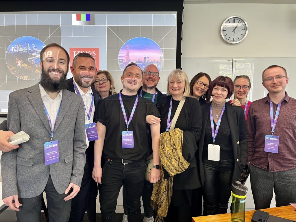 A group of 10 people from the PSM-AP team standing. They all are smiling and have purple conference badges hanging from their necks. They are in a small conference room infront of a projection, with some faces having lines projected on them.