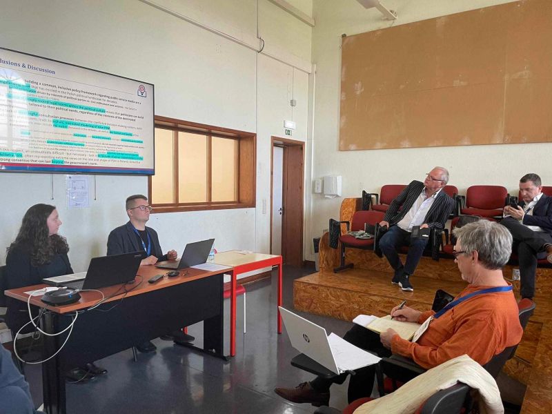 Photograph of Dagmara Furman and Filip Świtkowski presenting at the RIPE conference. They are seated infront of a projection showing unidentifiable highlighted text. Three audience members are also in the photograph, sitting in tierred seating.