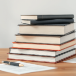 A stack of academic books with a pen and paper arranged studiously on a minimalist desk.